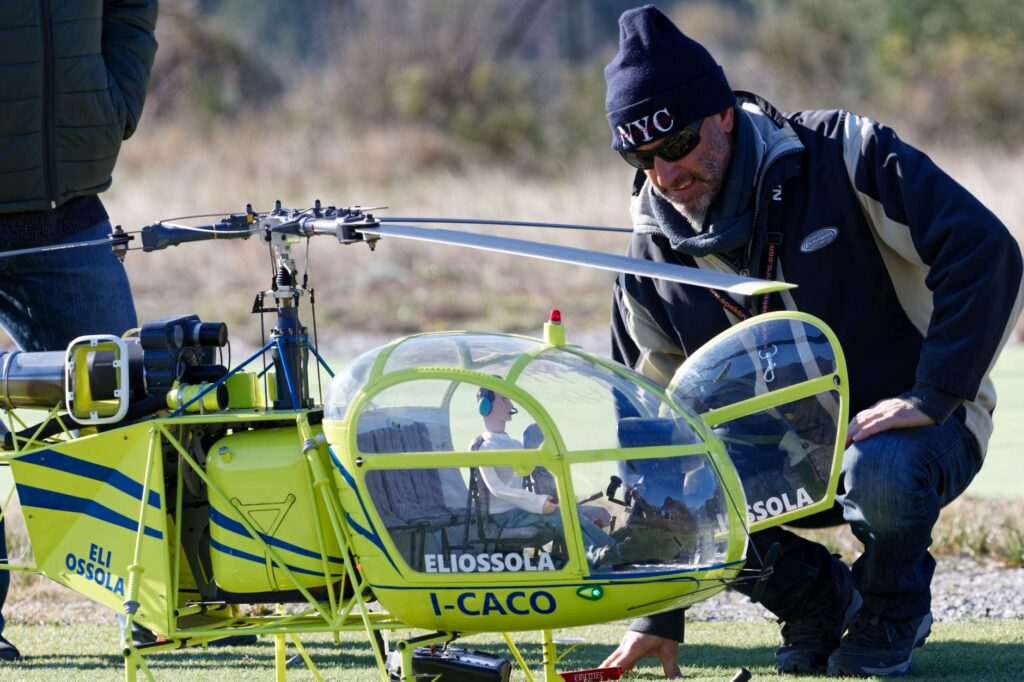 A man crouches beside a brightly colored model helicopter during an outdoor demonstration.