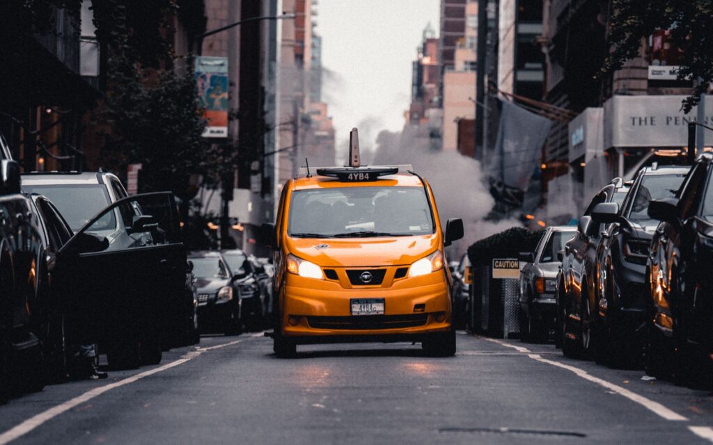 Yellow taxi cab navigates a busy New York City street with buildings and steam.