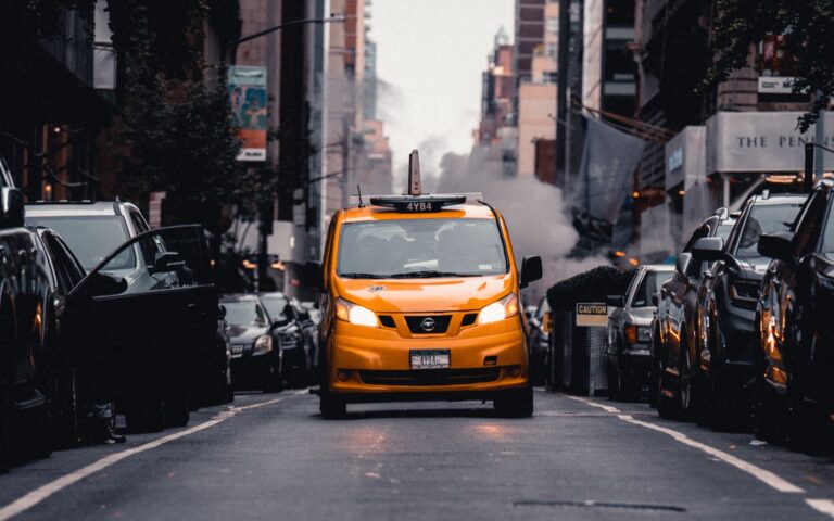Yellow taxi cab navigates a busy New York City street with buildings and steam.