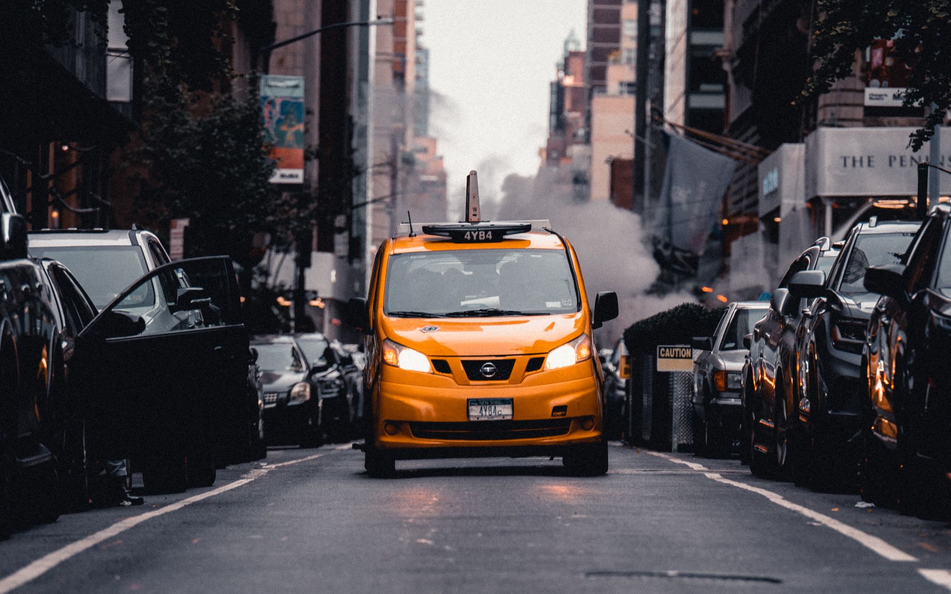 Yellow taxi cab navigates a busy New York City street with buildings and steam.