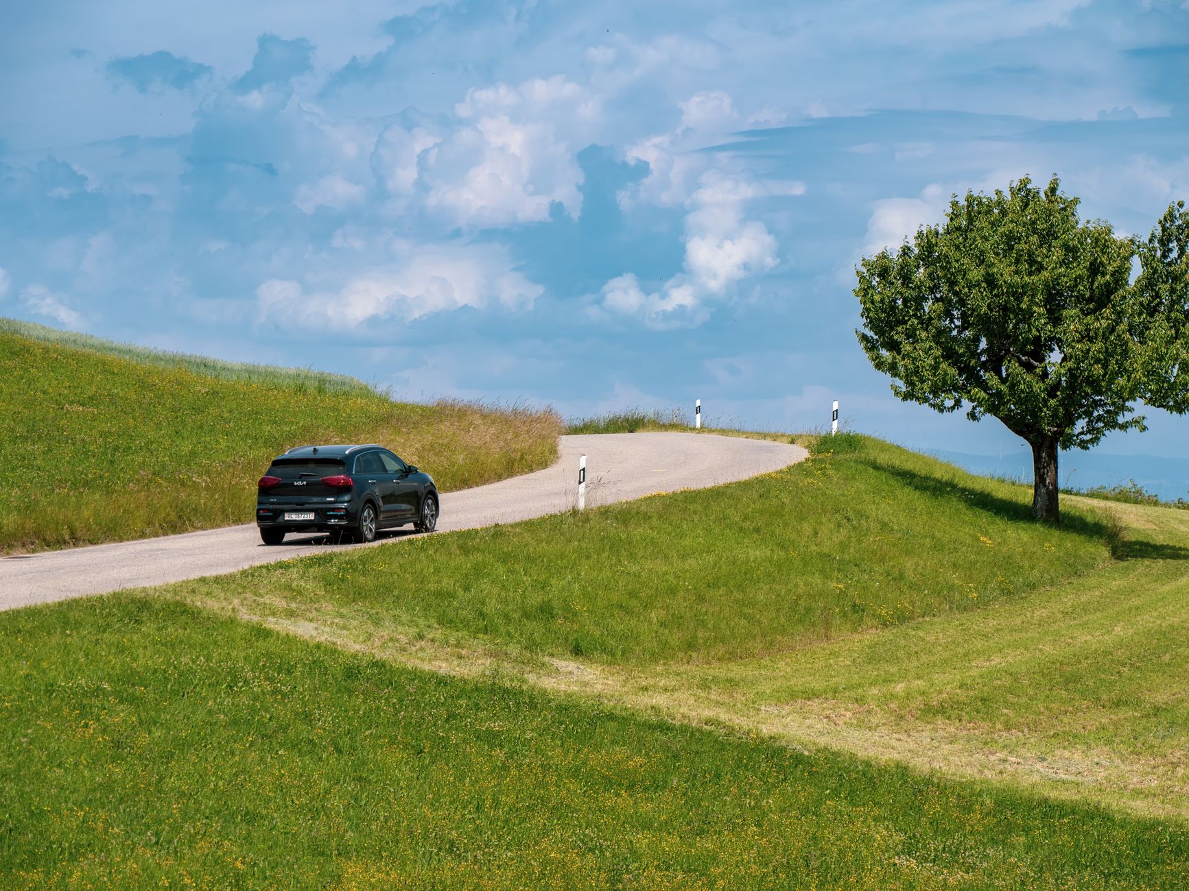 Black car cruising on a winding rural road under a blue sky with scattered clouds.