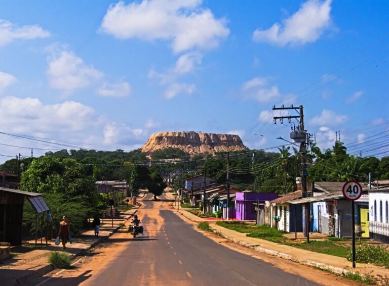 A vibrant town road with power lines leads to a majestic mountain under a blue sky.
