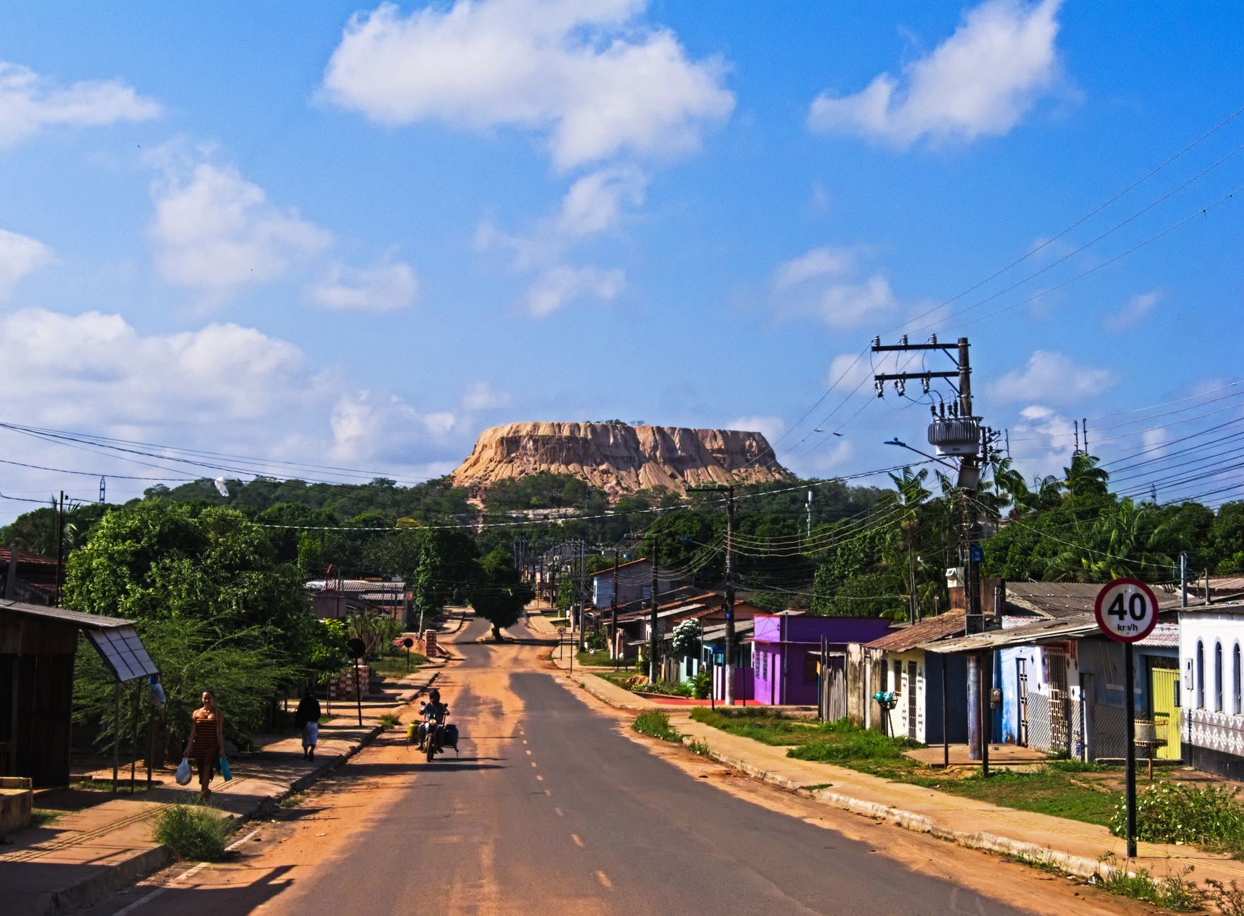 A vibrant town road with power lines leads to a majestic mountain under a blue sky.