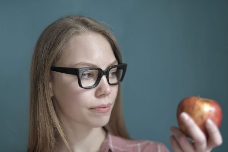 Woman wearing glasses holding an apple, conveying a thoughtful mood indoors.