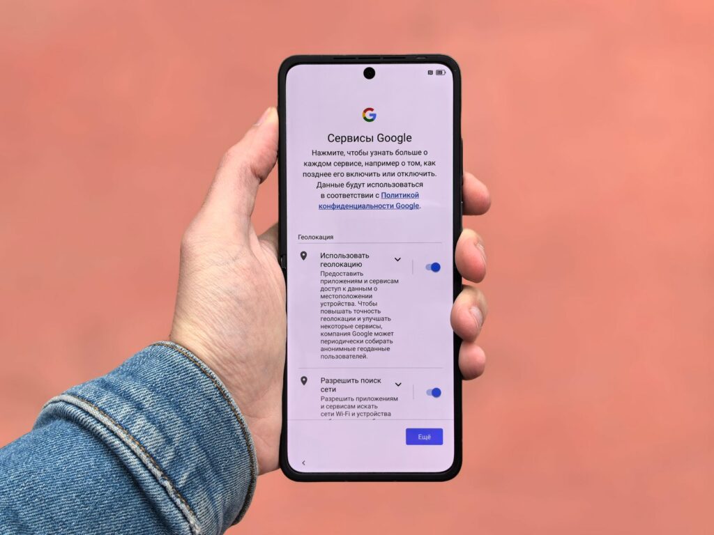 A person holding a smartphone displaying Google settings on a simple background.